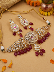 Gold-toned traditional Indian choker jewelry set featuring a necklace and matching earrings adorned with clear polki stones and deep red bead drops, arranged on a beige surface with decorative orange flower petals and a copper pot in the background.