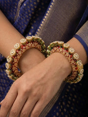 A close-up of a person wearing ornate traditional Indian bangles on both wrists. The bangles are gold-plated with intricate red enamel and kundan pearls, worn over a blue patterned saree.