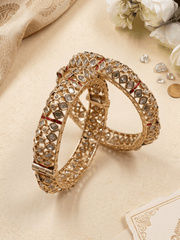 Two ornate gold-toned polki-style bangles decorated with clear stones and small red inlays, arranged on a beige surface with decorative fabric, flowers, and stones around them. Perfect for wedding, festive, and bridal wear.