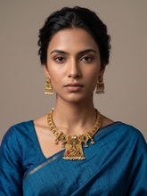 A woman wearing an ornate gold-plated antique gold short necklace and matching earrings with green and red stone accents, paired with a blue saree, against a plain beige background.