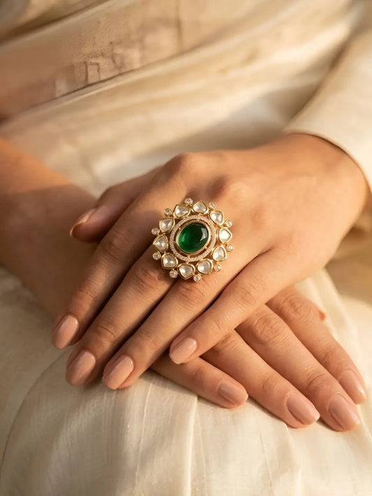 A close-up of a person's hands resting on cream-colored fabric, showcasing a large floral-design kundan ring with a central green stone and surrounding clear polki stones.