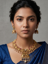 A woman wearing an ornate gold-plated traditional antique gold necklace with matching earrings and a blue sari, photographed against a plain gray studio background.