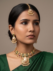 A woman wearing an ornate gold-toned traditional antique kundan jewelry set with a maang tikka, earrings, and a necklace, paired with a green saree, against a solid neutral background.