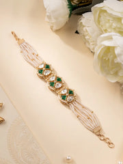 A gold-plated multi-strand pearl bracelet with green meena and clear polki stone embellishments, styled on a beige surface with white flowers.