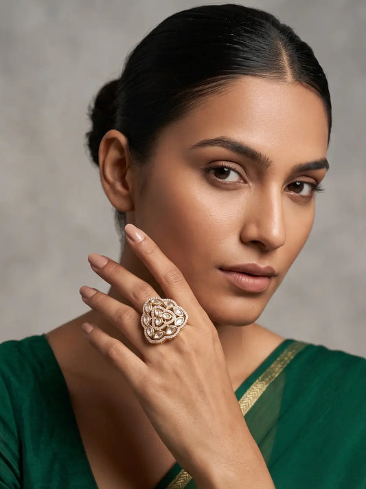 A close-up portrait of a woman wearing a large, ornate gold-plated kundan ring with a floral or paisley-like raised pattern on her finger, set against a soft gray studio background.
