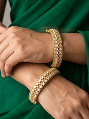 A close-up of a person's hands and arms wearing two gold-plated polki style bangles adorned with rows of white stones, set against a dark green fabric background.