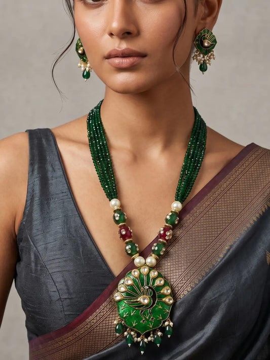 A close-up of a model wearing a traditional Indian jewelry set featuring a green beaded necklace with a large green and gold peacock pendant, matching green earrings, and a dark grey saree.