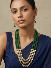 A woman wearing a multi-strand green beads and a kundan necklace with matching drop earrings, styled with a navy blue sari against a neutral beige background.
