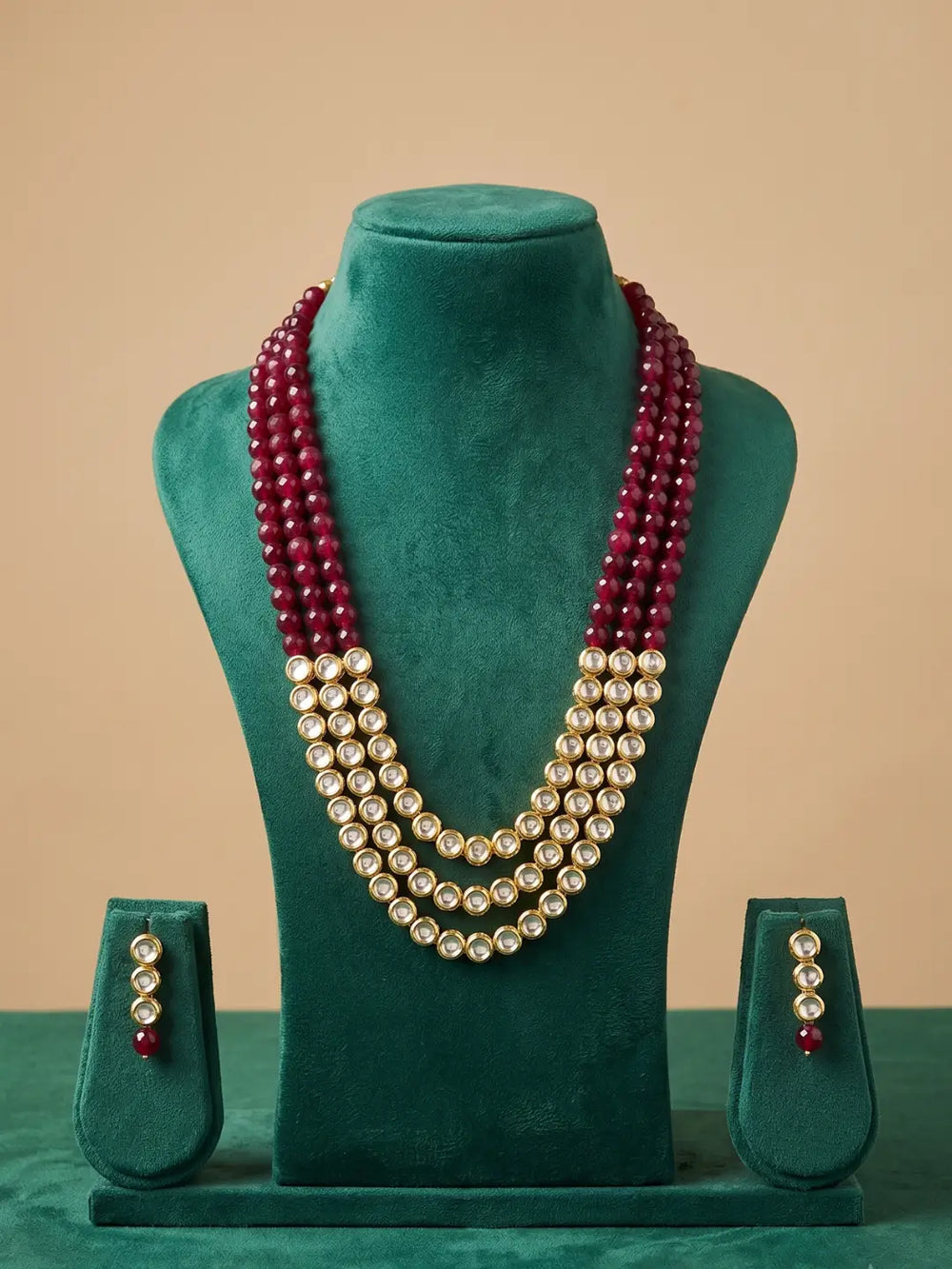 A three-layer re stones beaded kundan necklace set displayed on a green velvet mannequin bust, with matching drop earrings on small stands below.