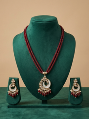 A traditional Indian jewelry set featuring a long red beaded necklace with a silver and white crescent mop pendant, accompanied by matching earrings, displayed on a green velvet mannequin bust.