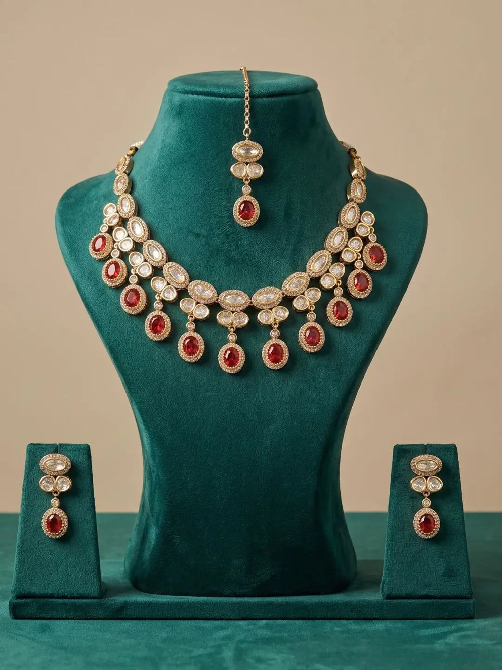 Gold-plated Indian jewelry set featuring a multi-layered necklace, matching pendant, and earrings, embellished with clear crystals and red gemstones, displayed on a teal velvet mannequin bust.