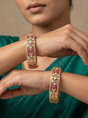 A close-up of a person wearing two ornate gold bangles decorated with red stones and clear stones, set against a soft neutral background and green clothing.