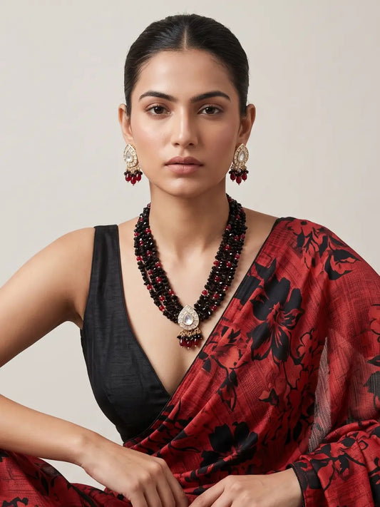 A woman wearing a red and black floral printed saree with a black sleeveless blouse, styled with a multi-strand red and black beaded necklace and matching earrings, set against a plain beige background.