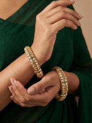 A close-up of a person wearing two ornate gold-plated green polki bangles adorned with green stones, paired with a dark green draped garment. Perfect for weddings, parties, and festive wear