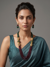 A woman wearing a multi-strand traditional long beaded necklace with deep red beads, gold-toned ornate links, and green accents, paired with matching drop earrings, against a solid gray studio background.