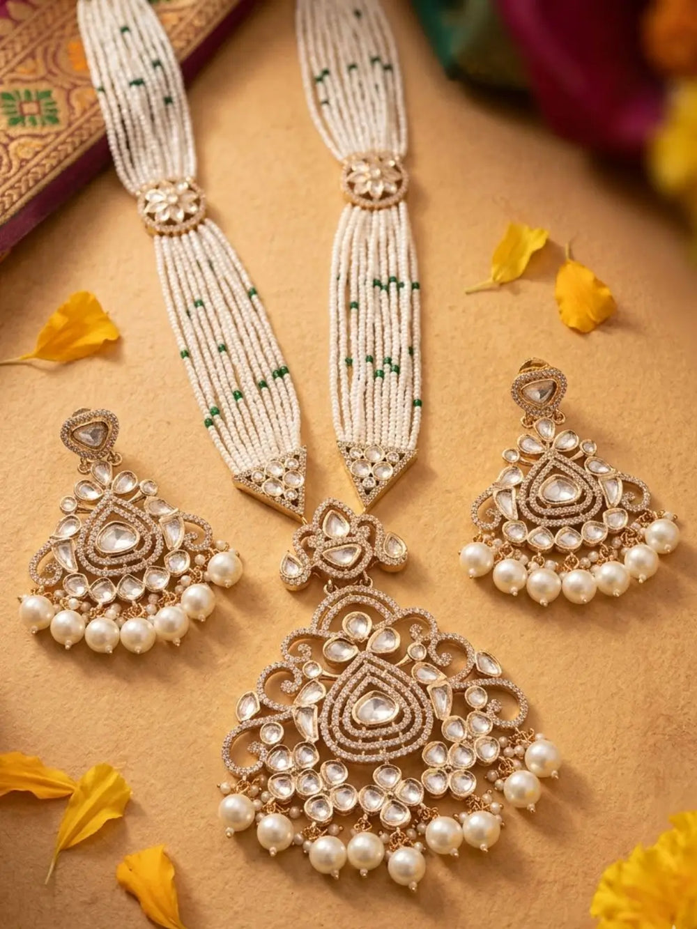 A traditional Indian bridal jewelry set featuring a gold-plated necklace and matching earrings adorned with white pearls, clear stones, and green bead accents, arranged on a warm tan background with scattered yellow flower petals.