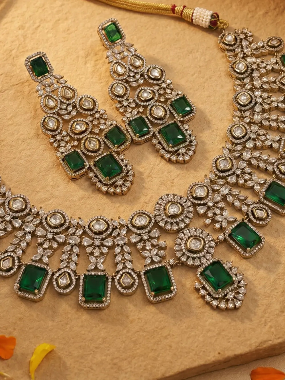 An ornate Indian ethnic jewelry set featuring a silver-tone necklace and matching earrings embellished with green rectangular emerald stones and round polki stones, displayed on a textured beige stone surface.