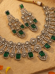 An ornate Indian ethnic jewelry set featuring a silver-tone necklace and matching earrings embellished with green rectangular emerald stones and round polki stones, displayed on a textured beige stone surface.