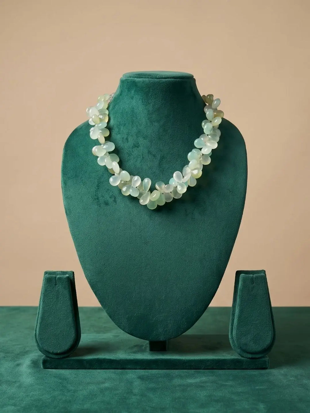 A beaded necklace made of pale iridescent shell-like beads displayed on a dark teal velvet jewelry bust against a warm beige background.