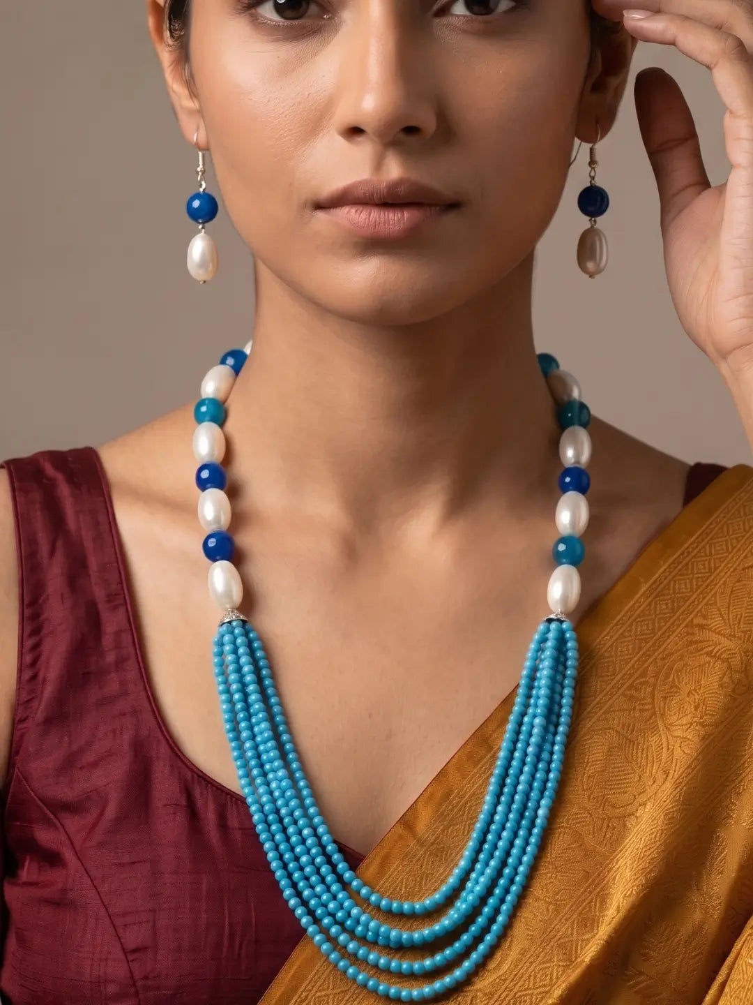 A close-up of a woman wearing a multi-strand turquoise and white pearl necklace, matching drop earrings, and a gold saree with a maroon blouse against a neutral beige background.