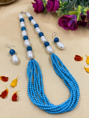 A blue and white beaded necklace set with matching drop earrings, displayed on a beige fabric background with decorative purple flowers and scattered petals.