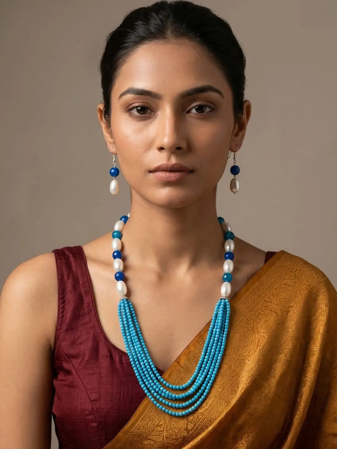 A woman wearing a multi-strand turquoise and white beaded necklace with matching drop earrings, styled with a maroon blouse and a gold saree against a neutral beige background.