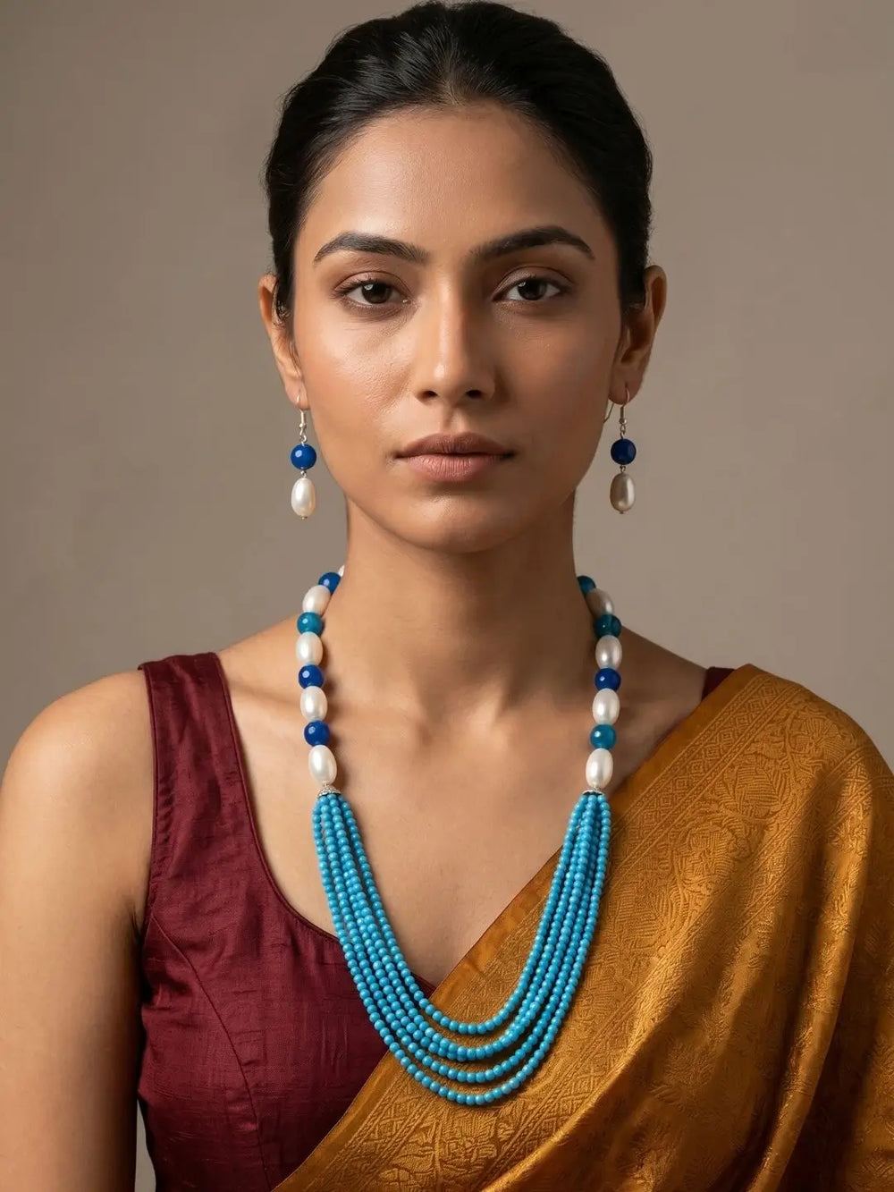 A woman wearing a multi-strand turquoise and white beaded necklace with matching drop earrings, styled with a maroon blouse and a gold saree against a neutral beige background.