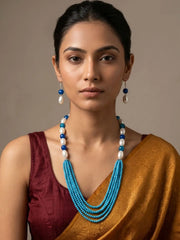A woman wearing a multi-strand turquoise and white beaded necklace with matching drop earrings, styled with a maroon blouse and a gold saree against a neutral beige background.