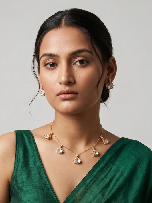 A woman wearing a delicate gold necklace with clear teardrop-shaped polki crystals and matching crystal stud earrings, paired with a green satin top, set against a plain light gray background.