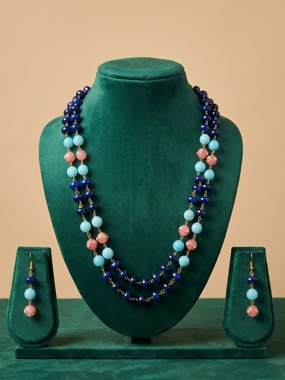 A beaded necklace and matching drop earrings displayed on a dark green velvet jewelry bust set against a warm beige background. The necklace features dark blue, light blue, and pink beads with small gold-toned spacers, and the earrings mirror this color scheme.