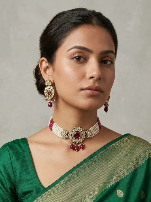 A woman wearing a green saree and matching jewelry, featuring a dual-tone mossanite polki choker necklace with red stones and matching drop earrings, set against a neutral gray background.