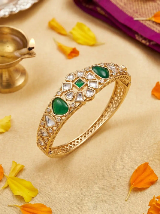 A gold-plated Kundan bangle bracelet decorated with green stone accents and clear polki work, displayed on a beige fabric background with scattered orange and yellow flower petals.