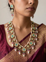 A close-up of a woman wearing an ornate gold-toned traditional necklace and matching drop earrings with mint green jadau and pink gemstones, paired with a deep purple silk saree.