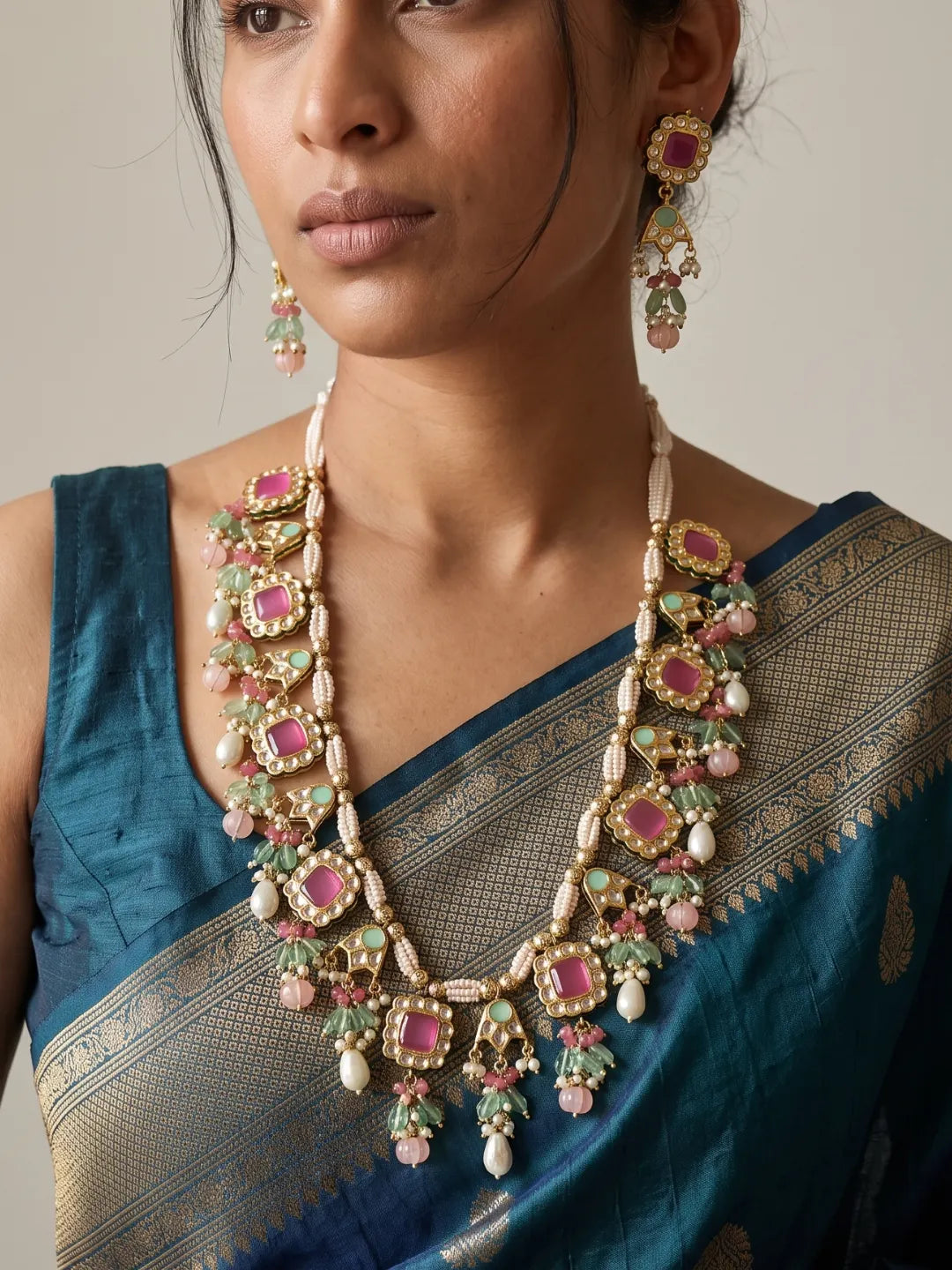 A close-up of a woman wearing an ornate gold-toned statement necklace and matching drop earrings with pink and green stones, paired with a teal blue saree.