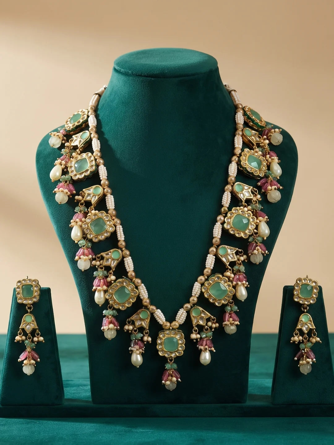 An ornate gold jewelry set displayed on a dark teal velvet mannequin bust, including a multi-colour necklace and matching drop earrings. The jewelry features mint green jadau stones, green enamel details, and white pearl-like beads, set against a solid beige background.