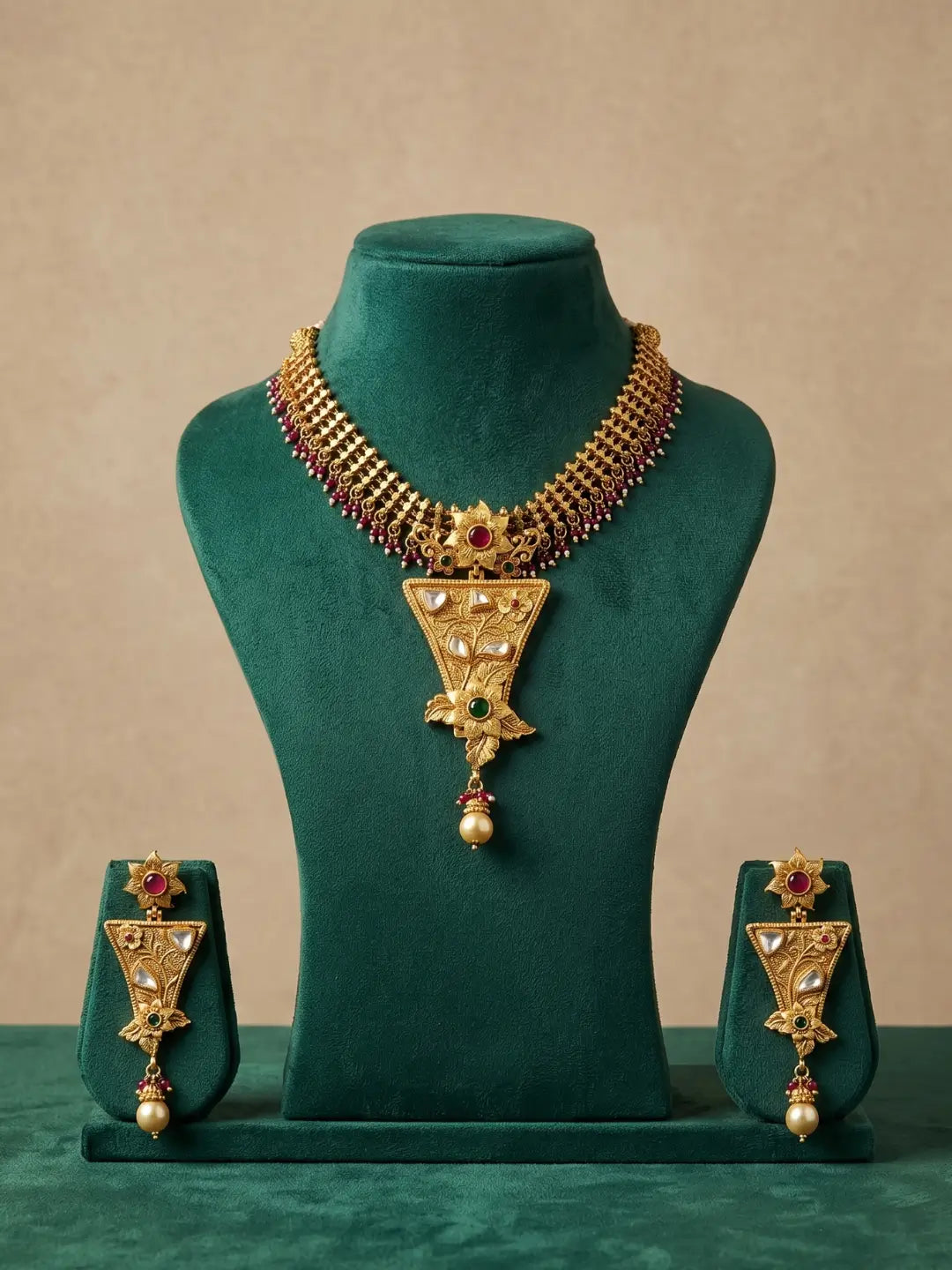 Gold-toned traditional jewelry set displayed on a dark green velvet mannequin bust, including a necklace with a large triangular pendant and matching earrings, set against a warm beige background.