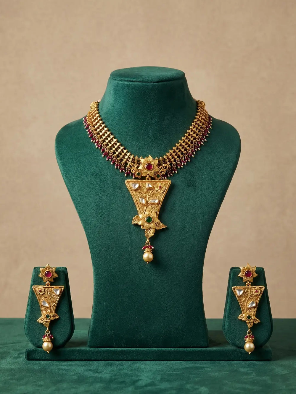 Gold-toned traditional jewelry set displayed on a dark green velvet mannequin bust, including a necklace with a large triangular pendant and matching earrings, set against a warm beige background.