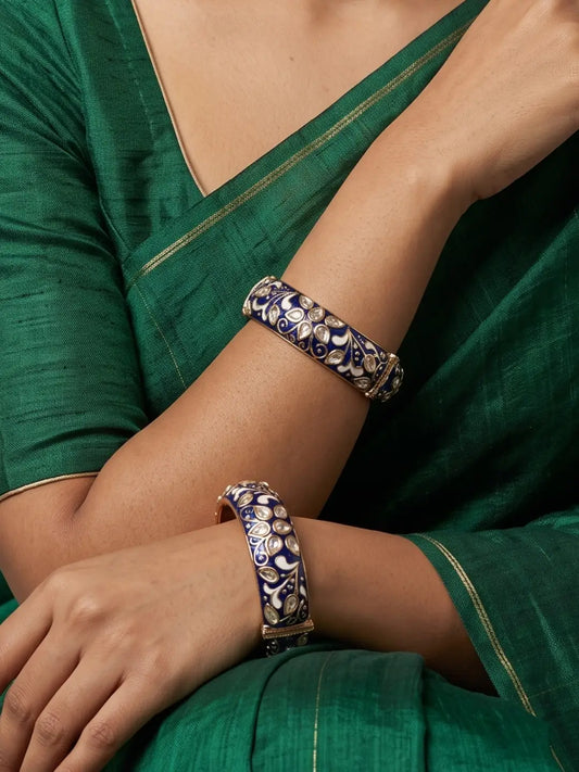 A close-up of a person wearing two ornate blue meenakari kundan bangles with blue, white, and gold floral patterns, paired with a green sari.