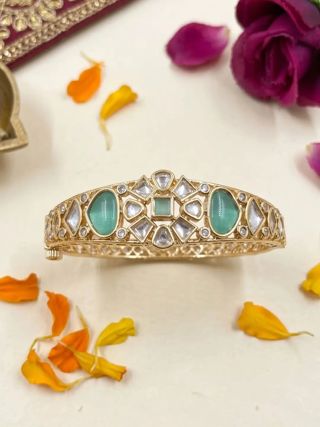 A gold-plated traditional Indian bangle bracelet studded with mint green stones and polki stones, displayed on a cream-colored surface with scattered orange and yellow flower petals.