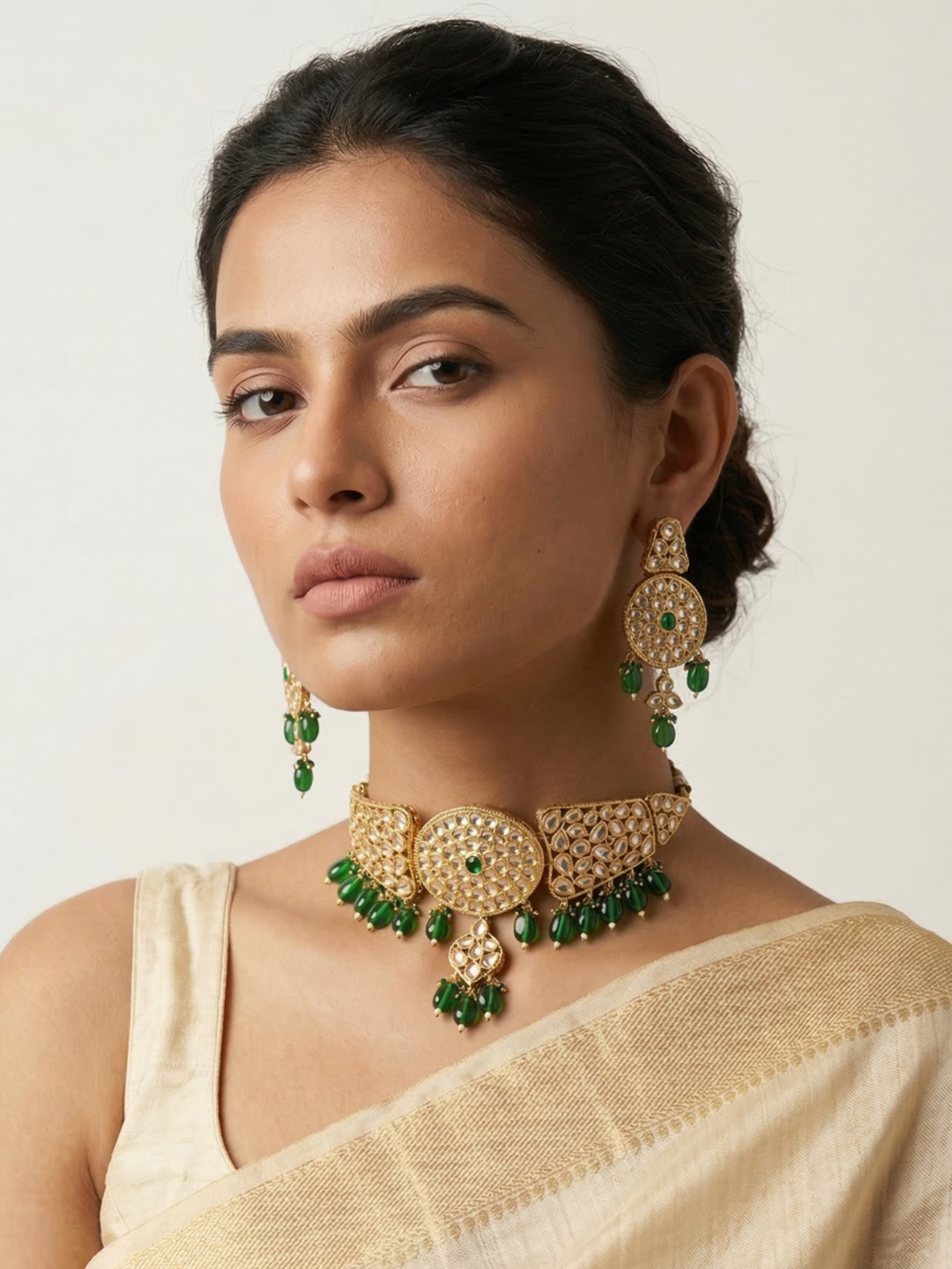 A woman wearing an ornate gold-toned choker necklace and matching drop earrings with green stone accents, paired with a light beige sari against a plain light background.