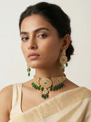 A woman wearing an ornate gold-toned choker necklace and matching drop earrings with green stone accents, paired with a light beige sari against a plain light background.