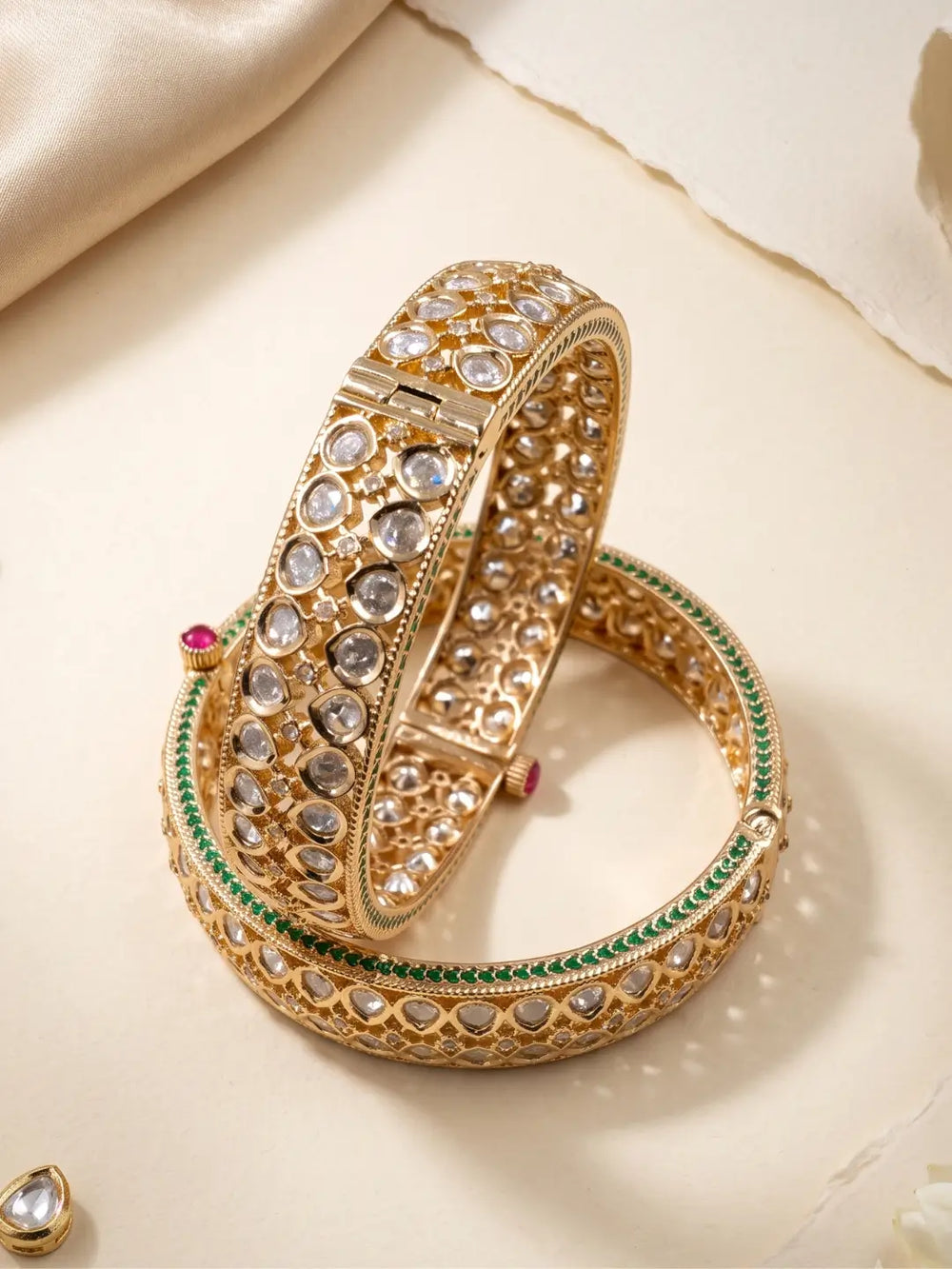 Two ornate gold-toned polki bangles with embedded clear stones, green enamel accents, and small red gem details, arranged on a cream satin surface.