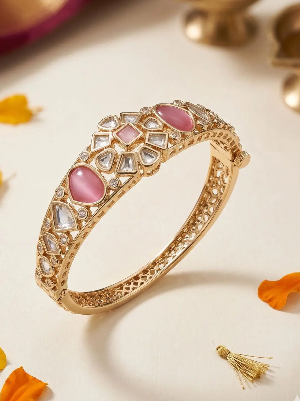 A gold-plated bangle bracelet with pink stone inlays and clear polki stones, displayed on a light cream surface with decorative orange petals and blurred brass elements in the background.