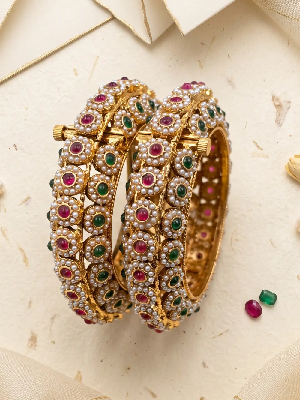 A pair of gold-plated multi-colour jadau bangles heavily embellished with white faux pearls, green emerald-cut stones, and pink ruby-cut stones, displayed on a textured beige background.