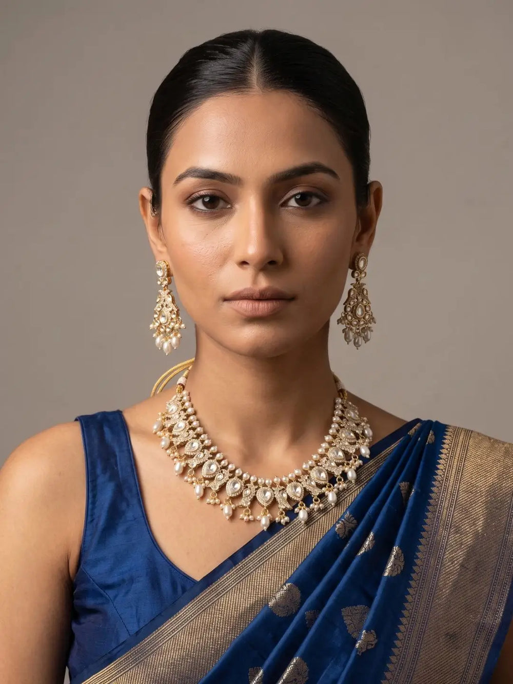A woman wearing a royal blue saree with a gold border and matching blue blouse, styled with a gold-plated peacock-design polki necklace set and matching earrings against a neutral gray background.