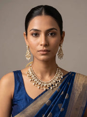 A woman wearing a royal blue saree with a gold border and matching blue blouse, styled with a gold-plated peacock-design polki necklace set and matching earrings against a neutral gray background.