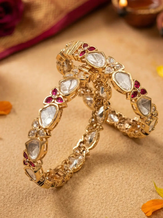 A pair of gold-plated Kundan bangles decorated with clear teardrop-shaped polki stones, small red floral accents, and clear round stones, displayed on a textured beige surface.