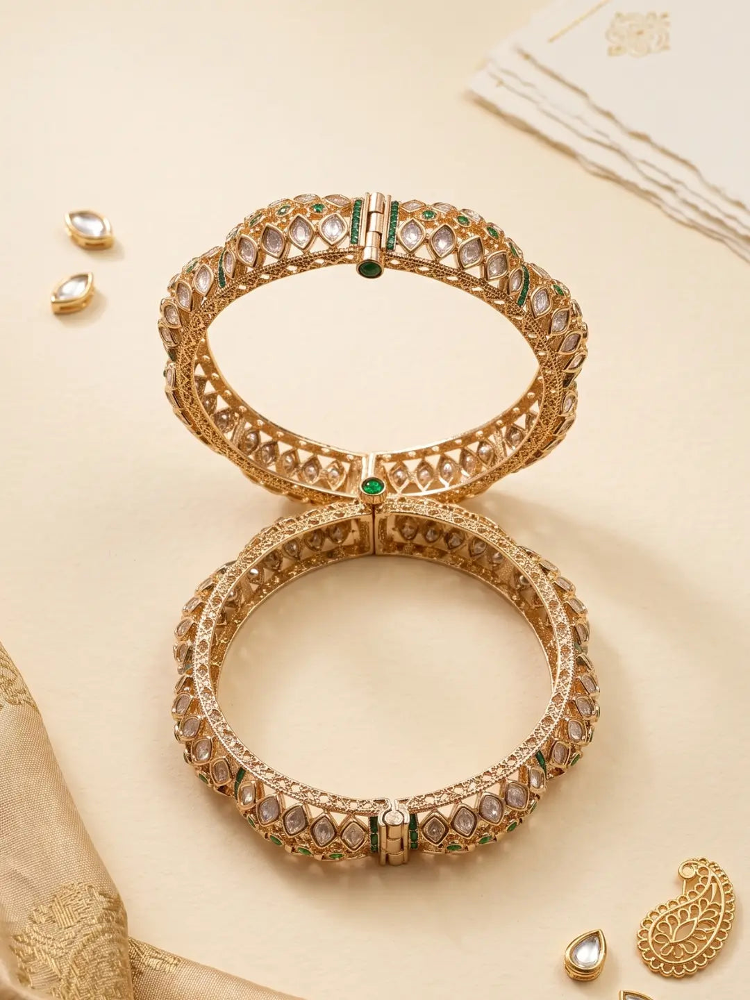 Two ornate gold-toned green polki kundan bangles with intricate gold-toned geometric patterns and small green stone accents, arranged on a warm beige background with decorative jewelry pieces and fabric.