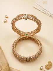 Two ornate gold-toned green polki kundan bangles with intricate gold-toned geometric patterns and small green stone accents, arranged on a warm beige background with decorative jewelry pieces and fabric.