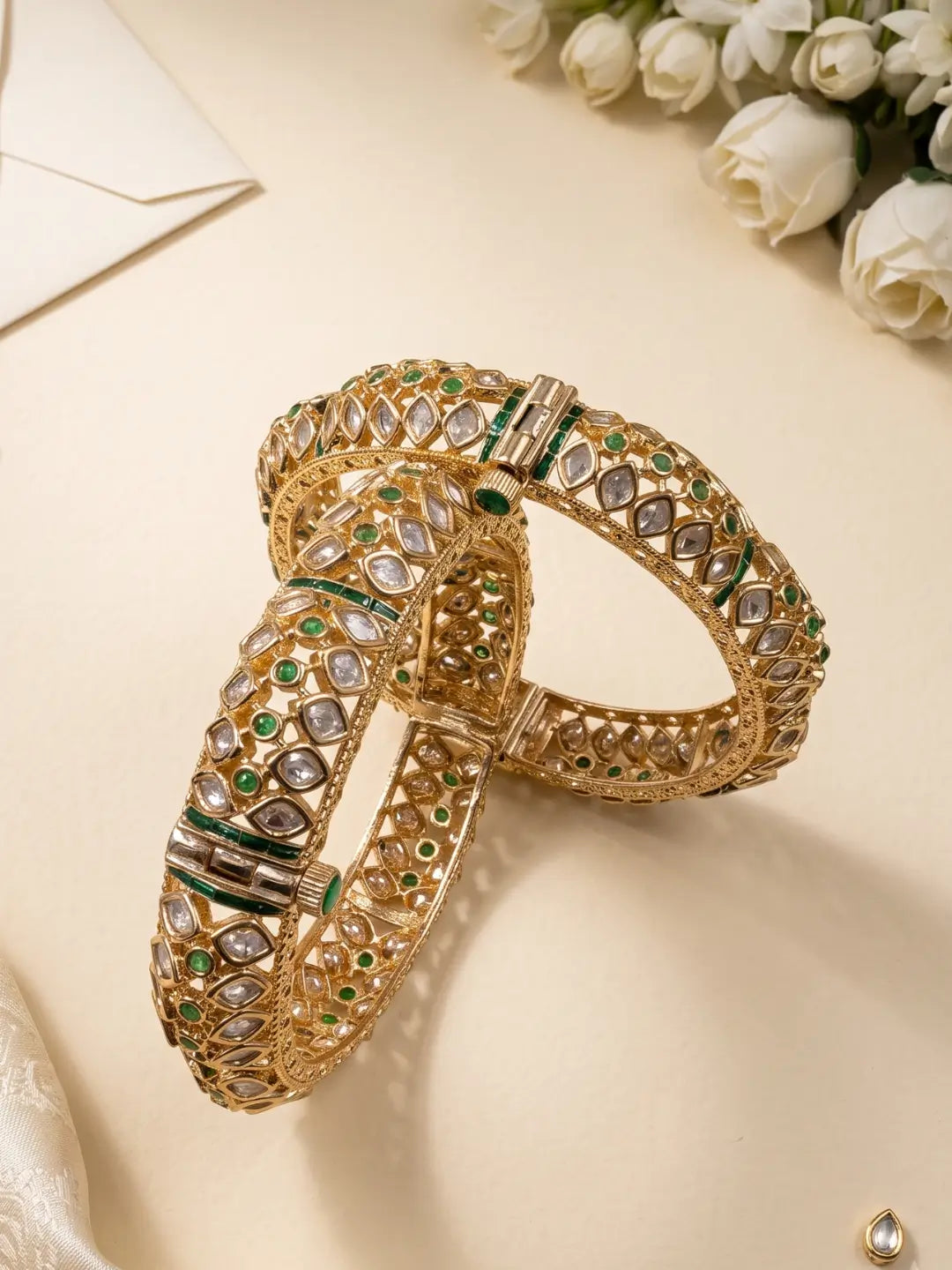 An ornate gold-plated uncut polki bangle bracelet decorated with polki stones and small green stones, on a light beige surface with white roses in the background.